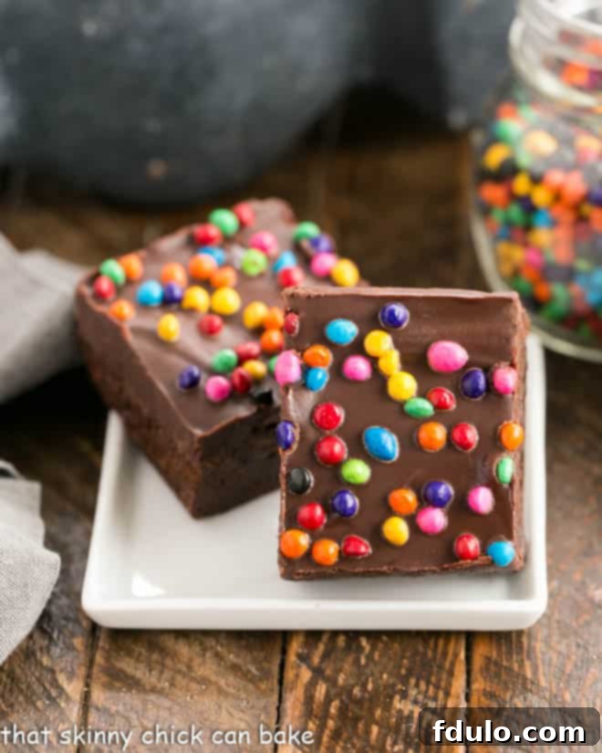 Two rich and fudgy Cosmic Brownies, topped with glossy ganache and rainbow sprinkles, presented on a small square ceramic plate.