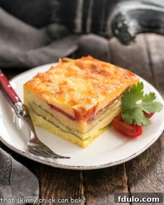 Low Carb Chile Rellenos Casserole - Layers of gooey cheese, eggs, tomatoes, and mild green chiles create an irresistible Tex-Mex egg casserole dish. A perfectly portioned slice of Low Carb Chile Rellenos Casserole on a white plate, garnished with fresh cherry tomatoes and a sprig of cilantro, highlighting its rich, cheesy layers and vibrant ingredients.