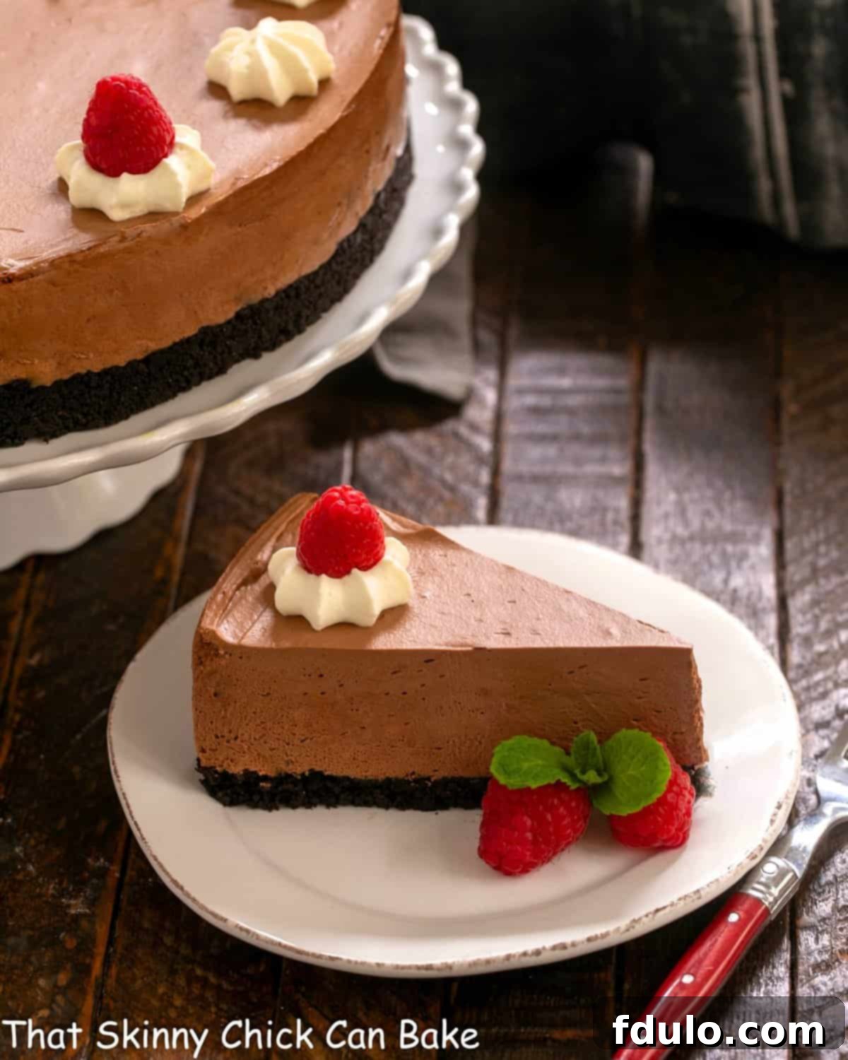A perfect slice of Chocolate Mousse Cake on a white dessert plate, with the full cake on a stand in the background. The slice shows the distinct crust and mousse layers, garnished with a single chocolate leaf.