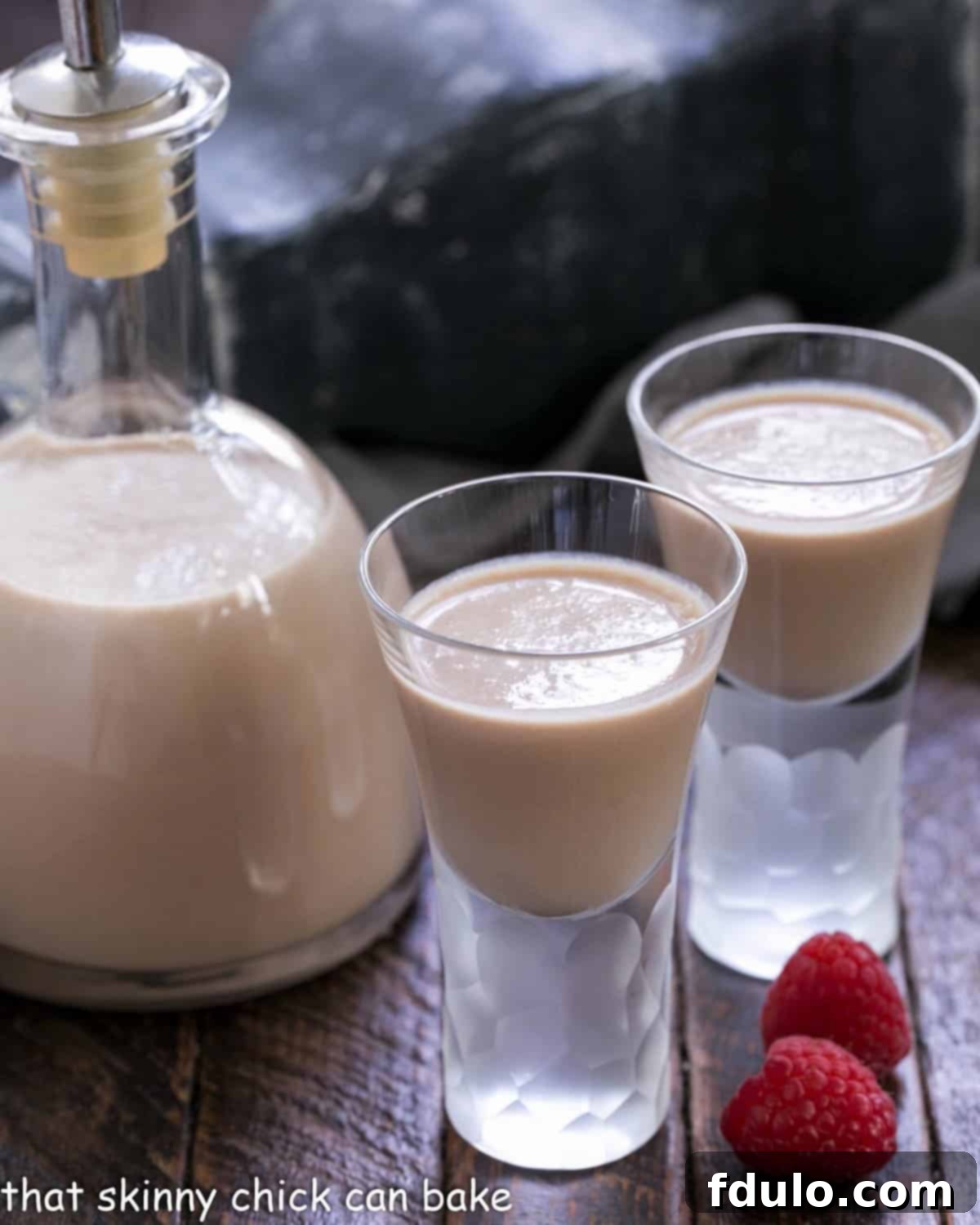 Irish cream recipe in a carafe and two shot glasses of Baileys with some fresh raspberries.