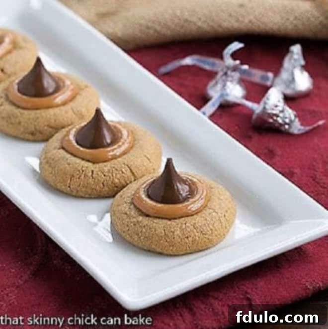 A festive white ceramic tray adorned with golden-brown Caramel Biscoff Blossoms Cookies, each topped with a glossy caramel center and a milk chocolate Hershey's Kiss. Perfect for holiday baking.