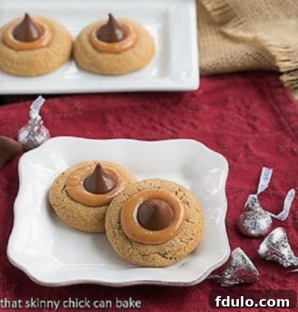 A close-up shot of several freshly baked Caramel Biscoff Blossoms resting on a square white plate, highlighting their golden edges, gooey caramel, and perfectly placed chocolate Kisses.