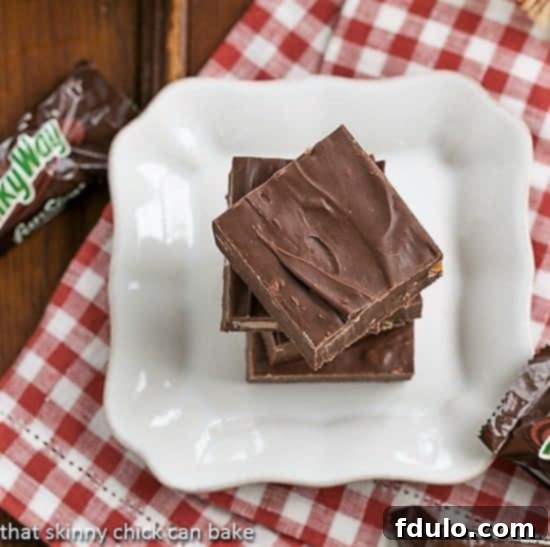 Close-up of freshly made Milky Way Fudge squares, neatly stacked on a white plate set against a festive red and white checkered napkin, highlighting their smooth texture.
