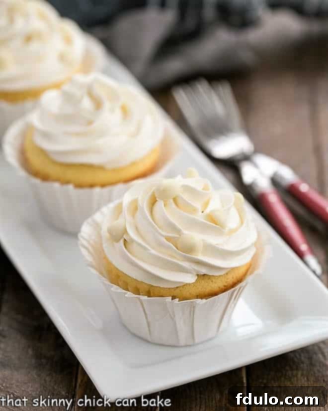 Ghirardelli Vanilyalı Cupcakeler, Beyaz Çikolatalı Swiss Meringue Kreması ile beyaz seramik bir tepside. Üzerleri beyaz çikolata rendesi ve minik taneciklerle süslenmiş.