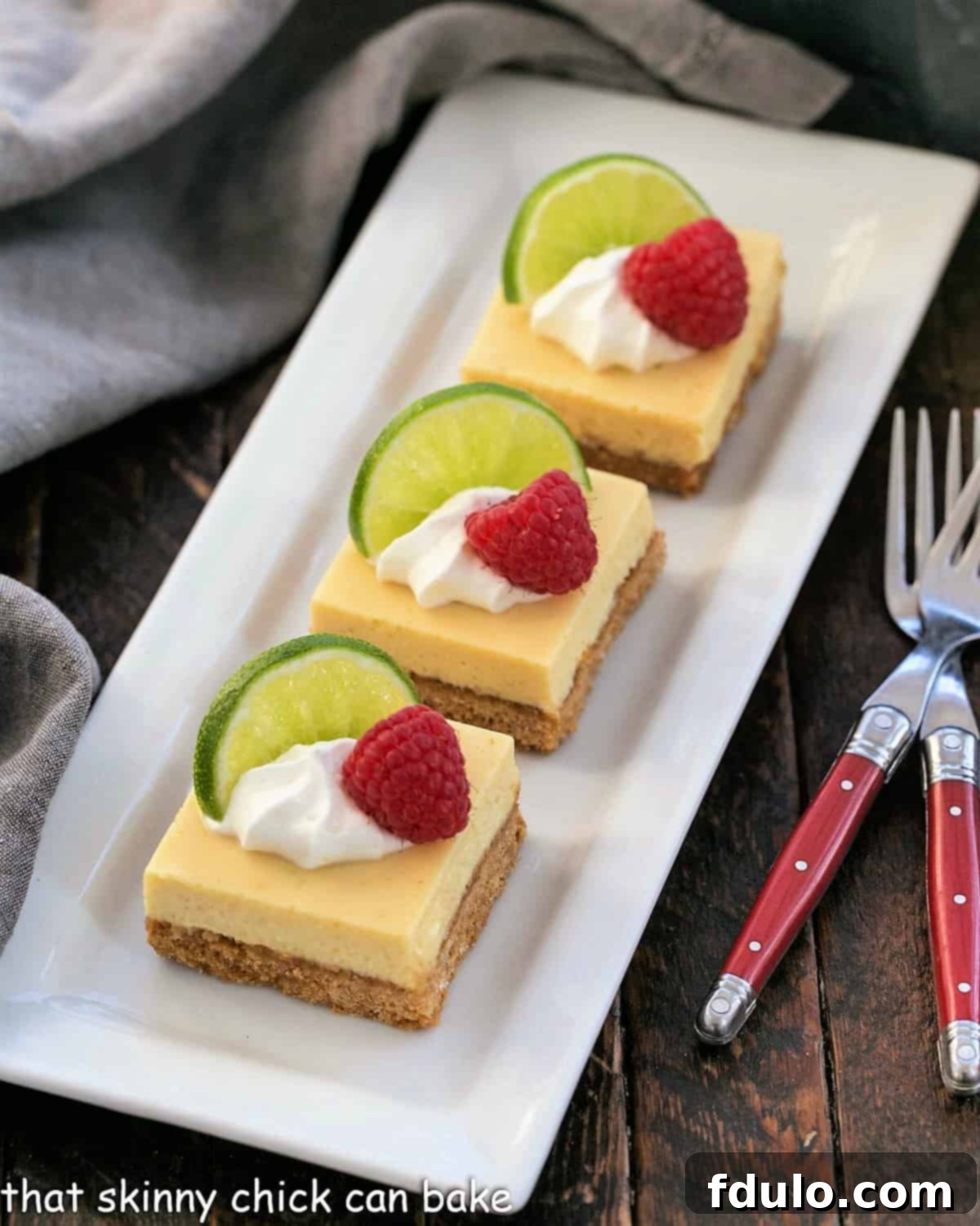 A close-up overhead shot of three Key Lime Pie Bars, artfully garnished on a white surface. Each bar is topped with a generous swirl of whipped cream, a delicate slice of fresh lime, and a vibrant scattering of fresh raspberries, creating an appealing contrast of colors and textures.