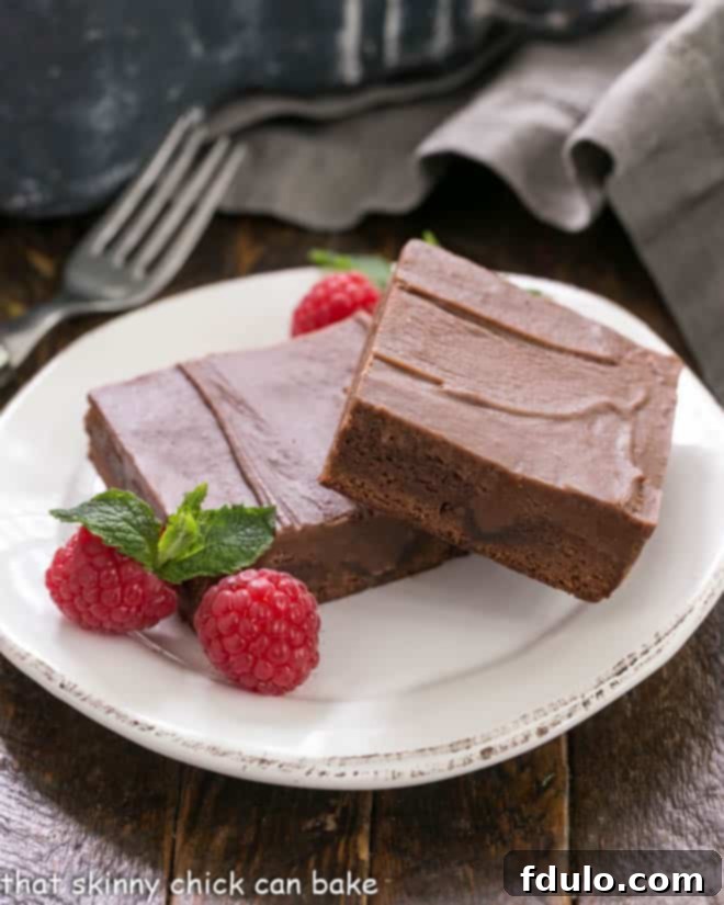 Chocolate Poke Brownies - The ganache seeps INTO the brownies!!! Two richly frosted chocolate poke brownies, garnished with fresh raspberries and a sprig of mint, presented on a pristine white dessert plate.