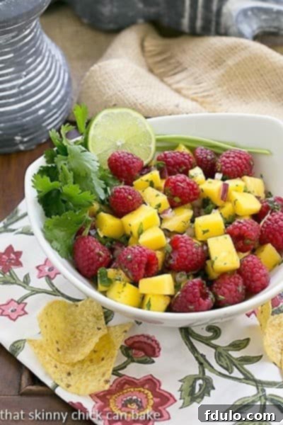 A close-up shot of Raspberry Mango Salsa in a pristine white bowl, garnished with a sprig of cilantro and a lime wedge.