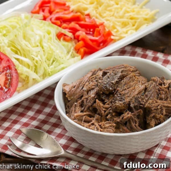 Close-up of Slow Cooker Beef Barbacoa served in a white bowl, garnished with fresh cilantro and lime wedges, surrounded by a variety of taco toppings.