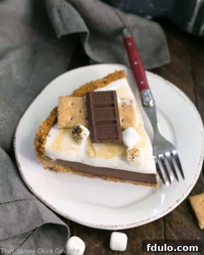 A perfectly sliced S'mores Pie on a white dessert plate, revealing its layers of graham cracker crust, chocolate filling, and fluffy toasted marshmallow topping.
