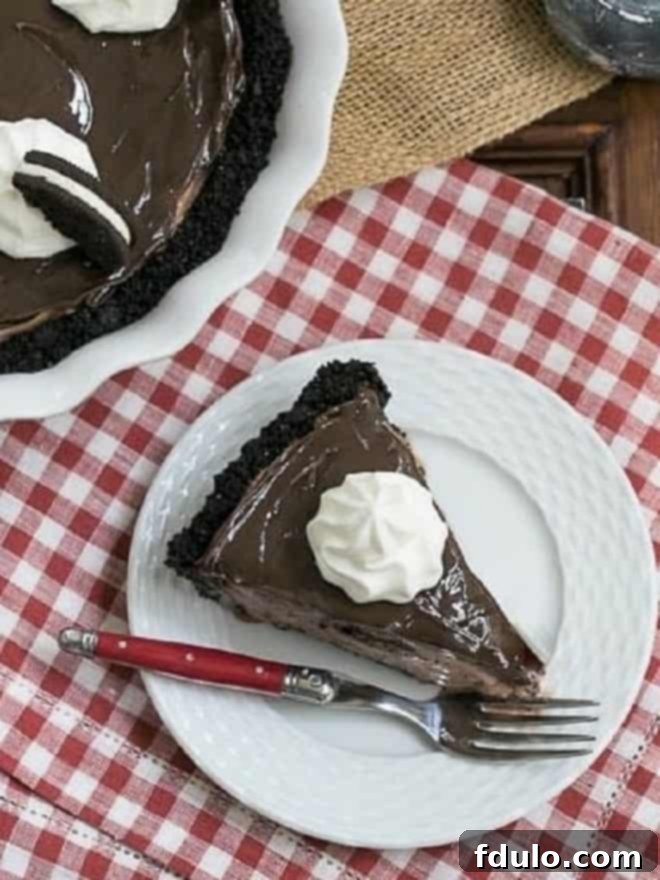 A perfectly sliced piece of Fudge Topped Chocolate Ice Cream Pie, showing its distinct layers of cookie crust, chocolate ice cream, and smooth fudge topping, on a white plate.