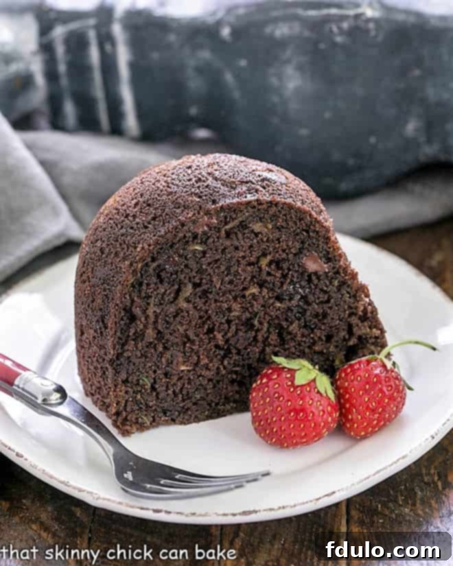 Chocolate Zucchini Cake - the addition of grated zucchini makes for a moist, delectable chocolate cake! A perfectly sliced piece of Chocolate Zucchini Bundt Cake on a white plate, accompanied by a red-handled fork and two fresh strawberries, highlighting its moist interior.