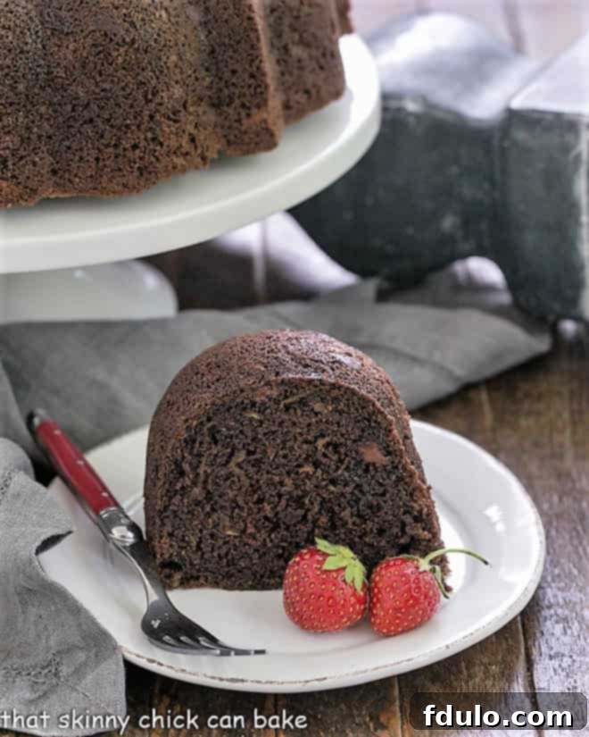 Chocolate Bundt Cake - the addition of grated zucchini makes for a moist, delectable chocolate cake! A beautiful slice of zucchini cake presented on a white plate, with the entire Chocolate Zucchini Bundt Cake visible on a white cake stand in the background.