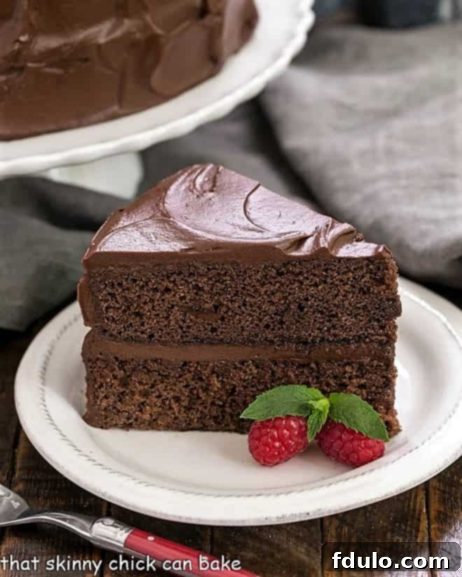 A perfect slice of Devil's Food Cake, showing its rich, dark interior and creamy chocolate frosting, served on a white dessert plate with an elegant red-handled fork, and a full cake on a stand blurred in the background.
