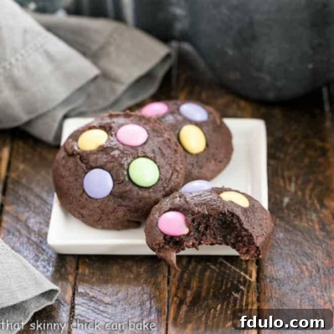 Rich, chewy Brownie Cookies filled with chocolate chips and topped with Easter M&Ms 3 Brownie Cookies on a square white plate, one with a bite removed, revealing the fudgy interior and chocolate chips.
