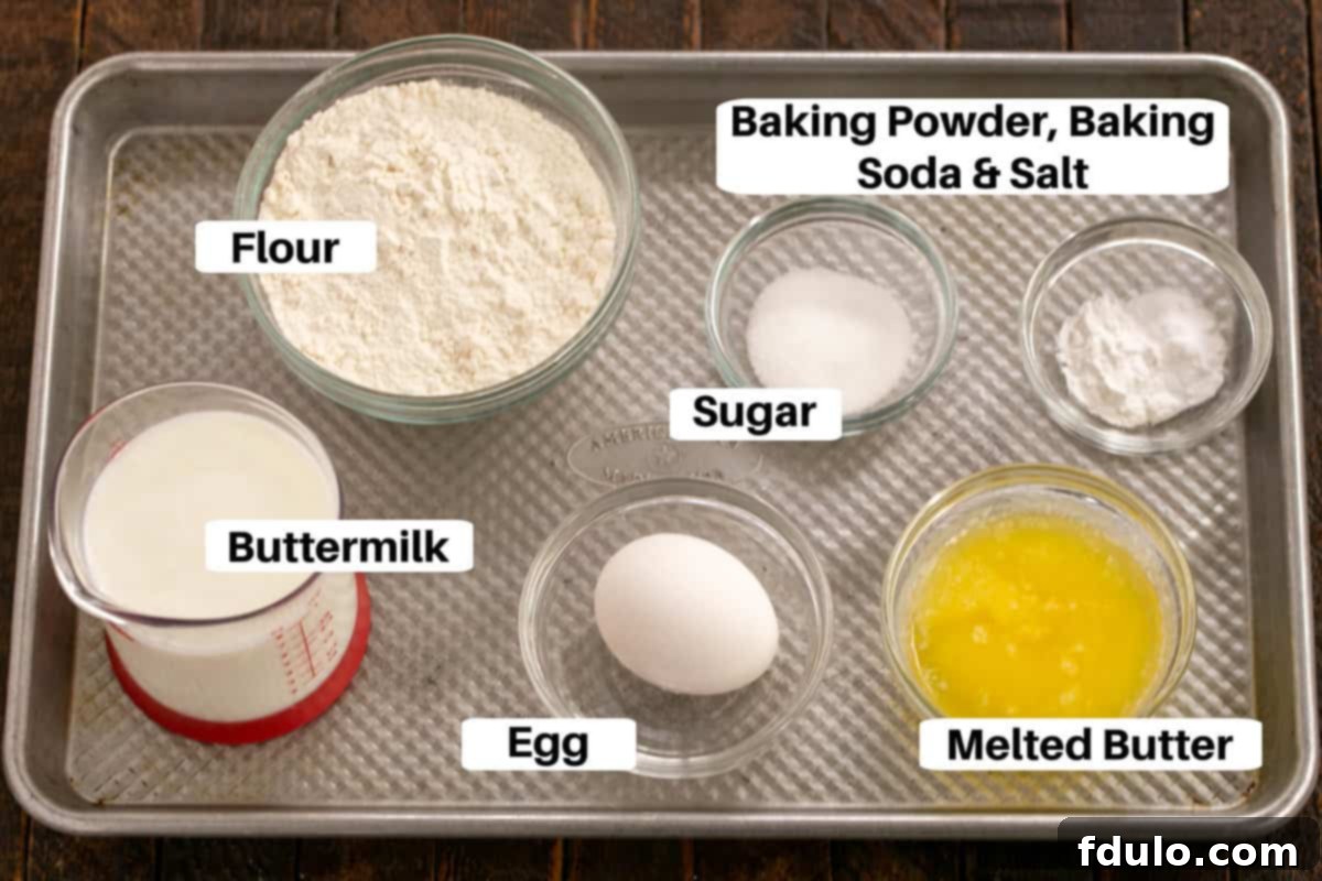 A close-up of various buttermilk pancake ingredients, including flour, sugar, baking soda, baking powder, eggs, butter, and a bottle of buttermilk, all neatly arranged on a metal sheet pan.