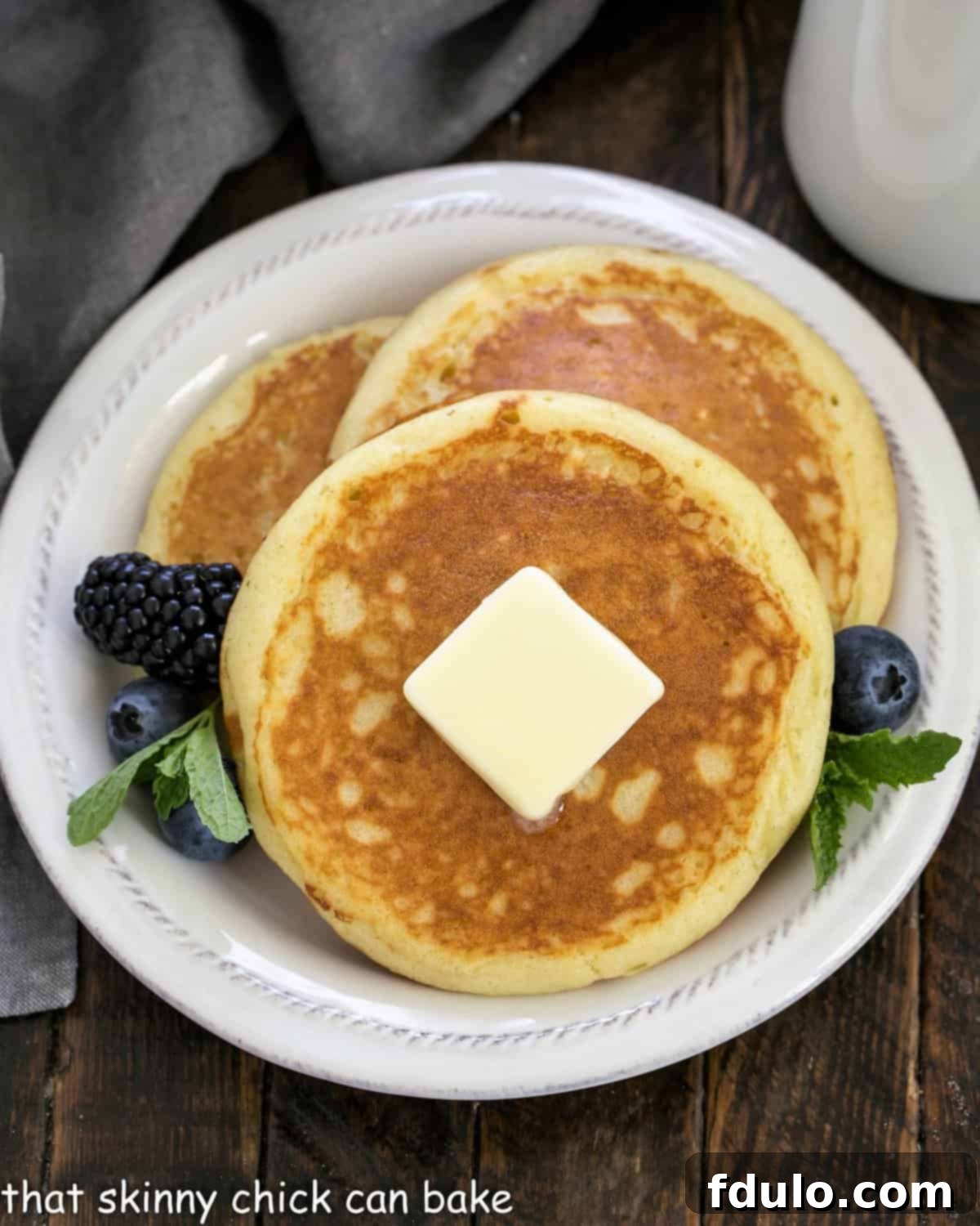 An overhead shot showcasing three beautifully cooked, fluffy buttermilk pancakes, artfully topped with a dollop of melting butter and garnished with vibrant berries and a sprig of fresh mint.