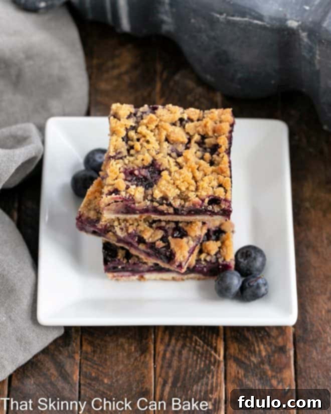 A stack of three perfectly cut blueberry streusel bars resting on a square white dessert plate, adorned with a scattering of fresh blueberries, ready to be enjoyed.