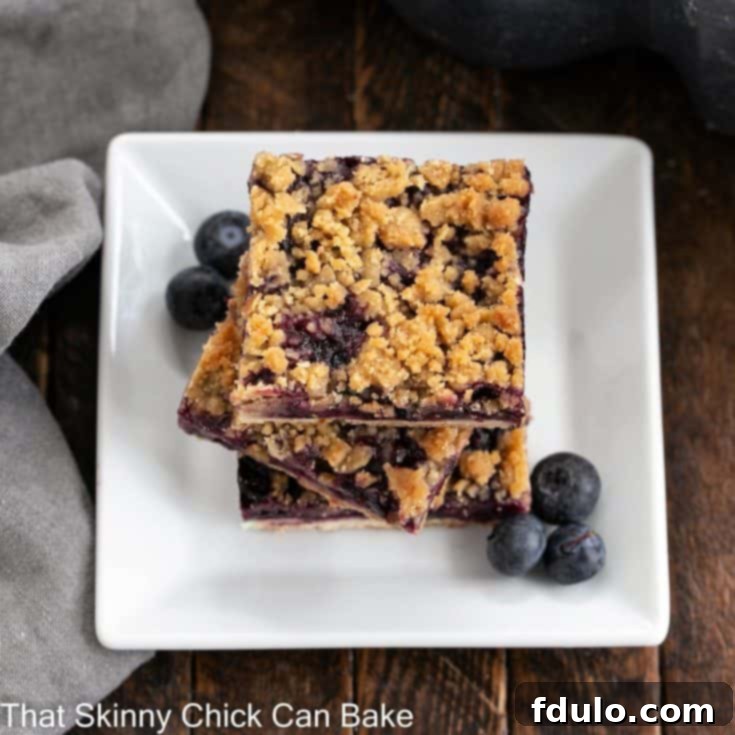 A beautifully composed stack of blueberry streusel bars on a pristine white ceramic plate, perfectly showcasing the golden streusel and a hint of the juicy blueberry filling. Fresh blueberries are scattered as a garnish, enhancing its appeal.
