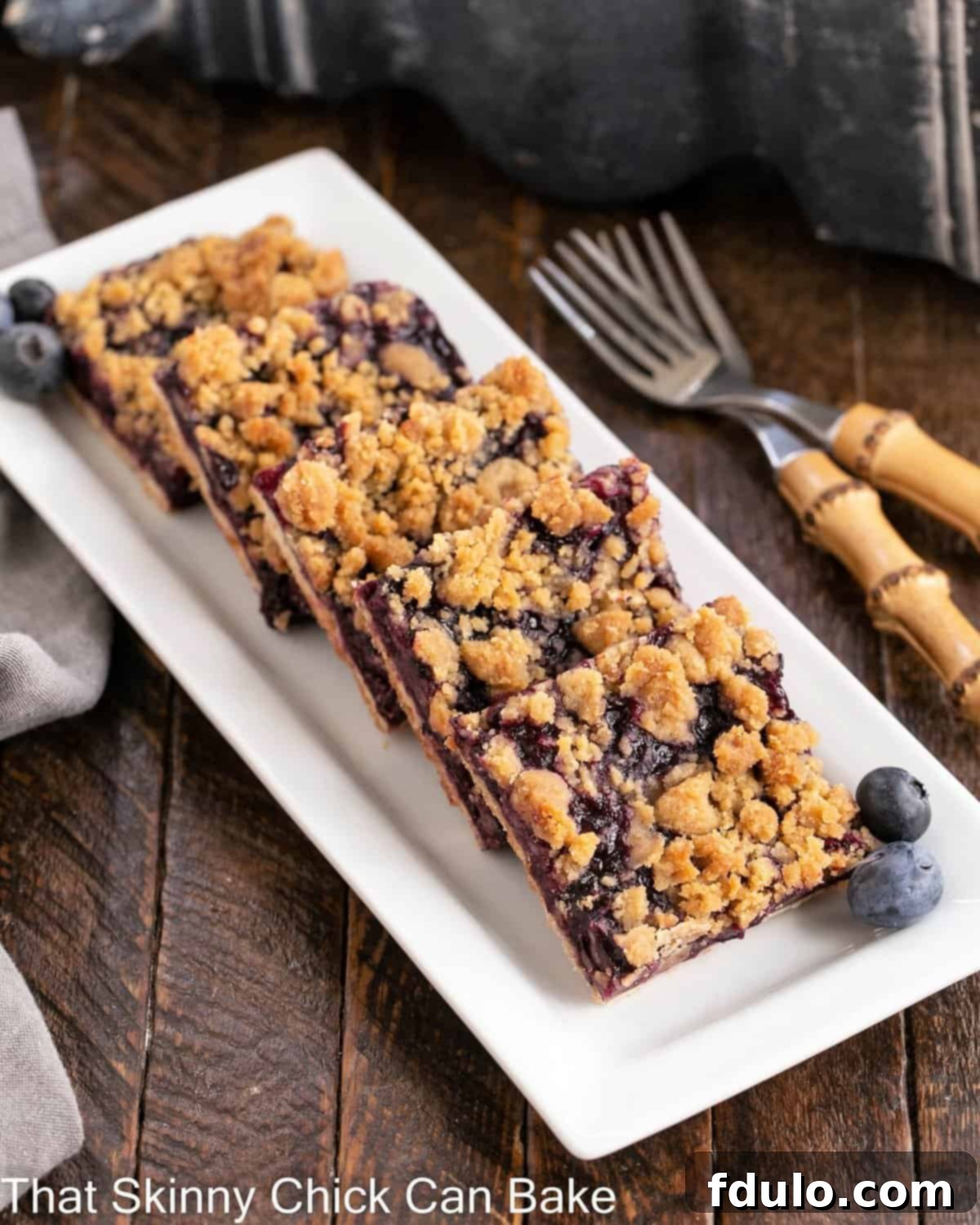 Close-up of golden-brown blueberry streusel bars neatly arranged on a pristine white serving tray, accompanied by two delicate bamboo forks, ready for enjoyment. The rich, crumbly streusel topping and vibrant blueberry filling are clearly visible.