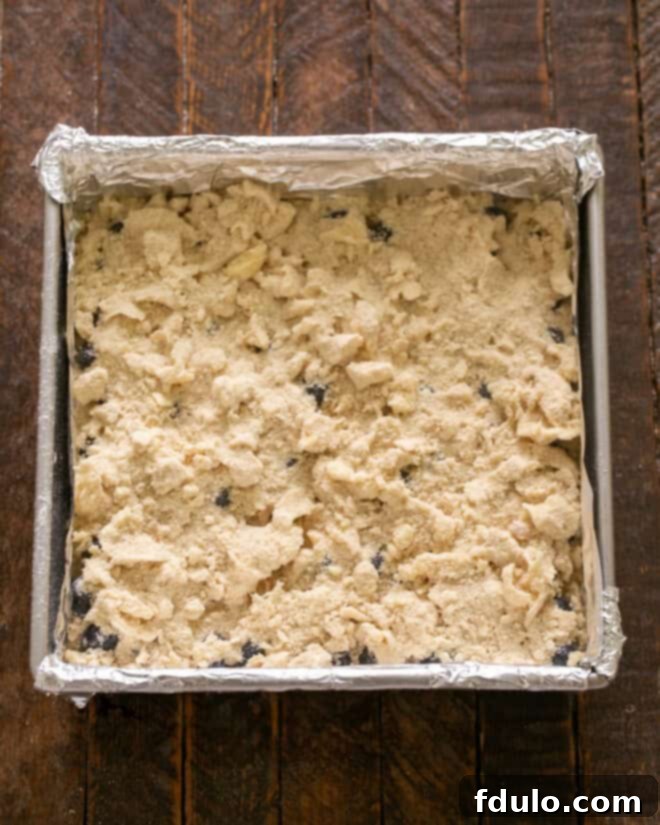 Step 6: Crumbling the buttery streusel topping evenly over the layer of fresh blueberries, creating a golden, textured blanket before baking.