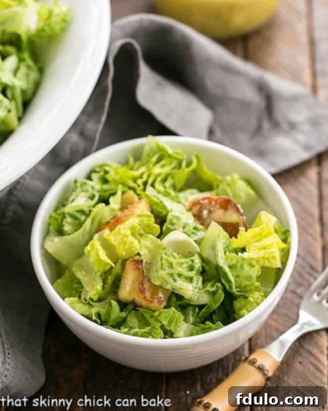 The Best Caesar Salad Dressing over romaine lettuce, croutons and Parmesan shavings - Make your favorite restaurant salad at home! Close-up view of a perfectly dressed Caesar Salad in a small white salad bowl with a bamboo-handled fork, showcasing crisp lettuce, golden croutons, and Parmesan shavings.