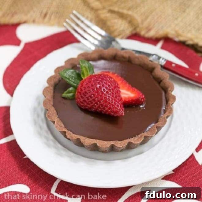 Exquisite individual Double Chocolate Tartlet topped with a fresh, vibrant red strawberry, resting on a pristine white plate, showcasing its rich brown crust and glossy chocolate ganache.