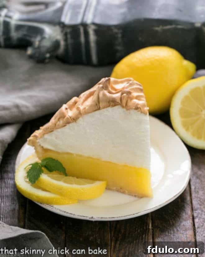 A beautifully presented slice of Mile-High Lemon Meringue Pie on a white dessert plate, garnished with fresh lemon slices and mint sprigs.