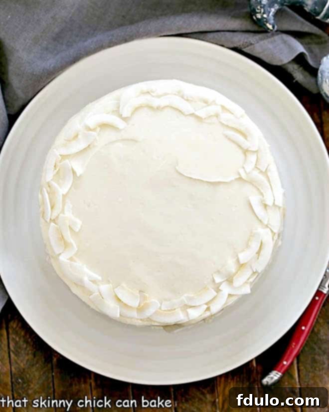 Overhead view of a beautifully decorated Coconut Cake on a white cake plate, garnished with coconut flakes.