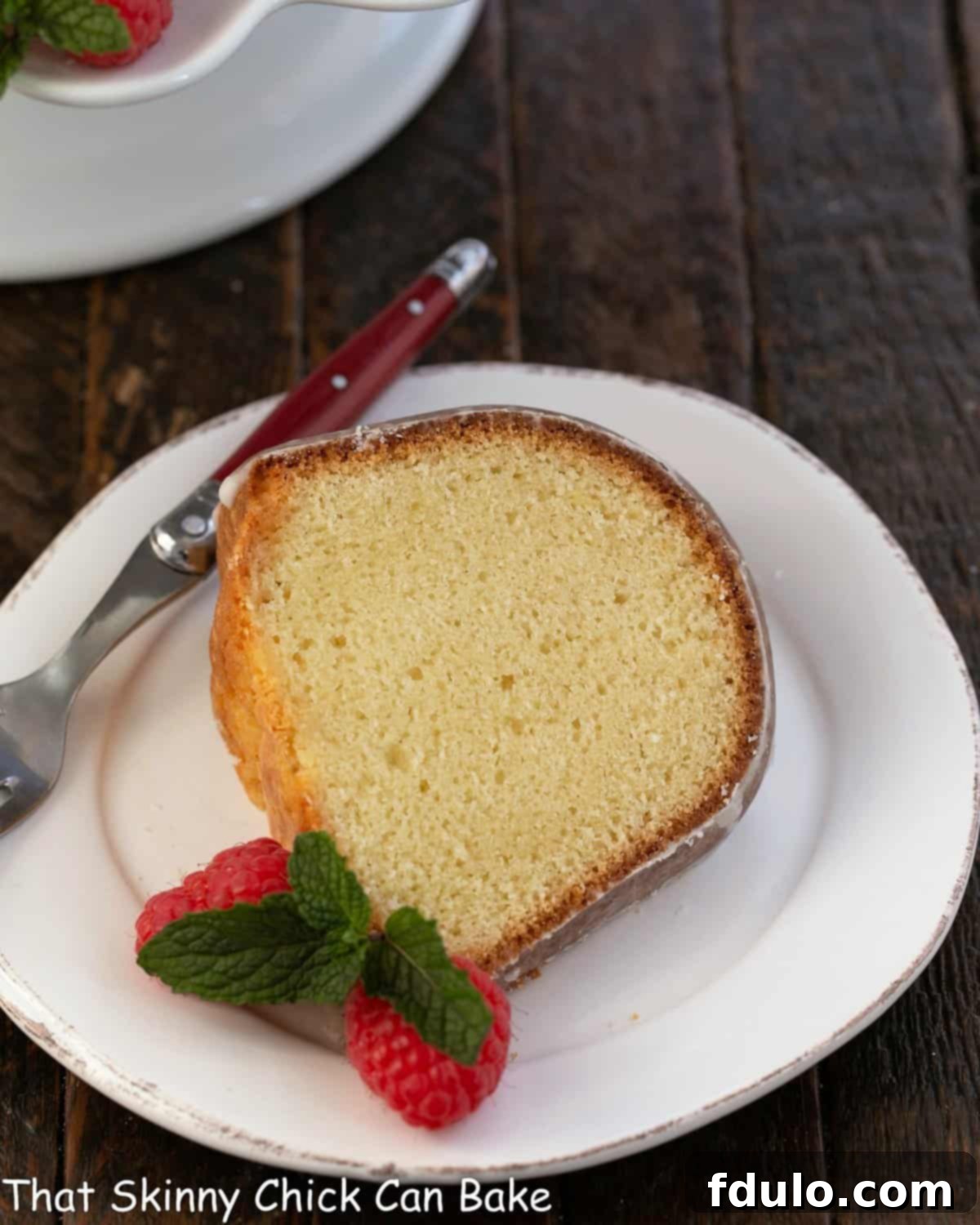 A slice of sour cream pound cake on a white desssert plate with a red handle fork and fresh raspberries.