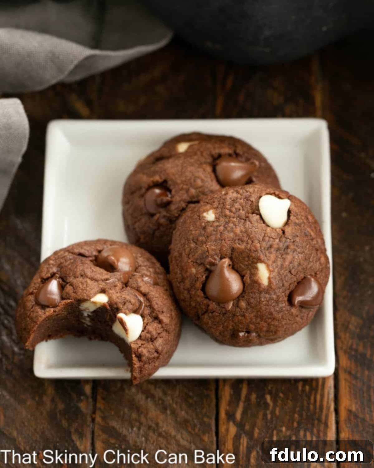3 chocolate chocolate chip cookies on a square white plate, one with a bite removed.