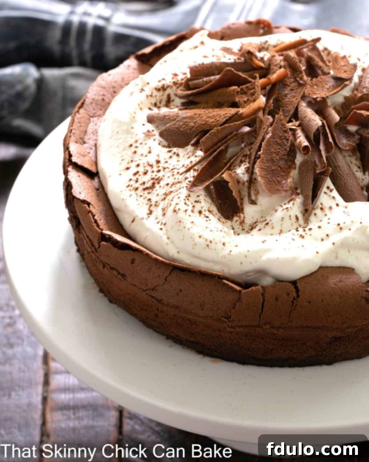 Chocolate Souffle Cake - Watch this cake rise and fall leaving a perfect well for sweet cream and berries Left side of a Chocolate souffle cake on a white cake stand, artfully garnished with whipped cream and chocolate shavings.