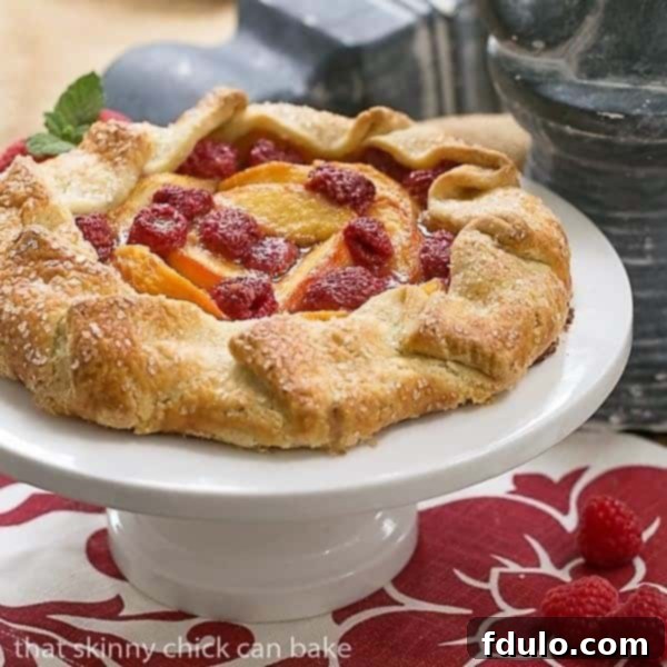 A slice of Peach Raspberry Galette served on a white ceramic plate, garnished with fresh raspberries and a sprig of mint, highlighting its inviting golden crust.