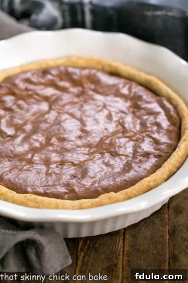 Fudgy Brownie Pie - Rich brownie filling studded with chocolate chips and baked in a flaky pastry shell! A whole Fudgy Brownie Pie sitting in a beautiful white, fluted ceramic pie plate on a wooden surface.