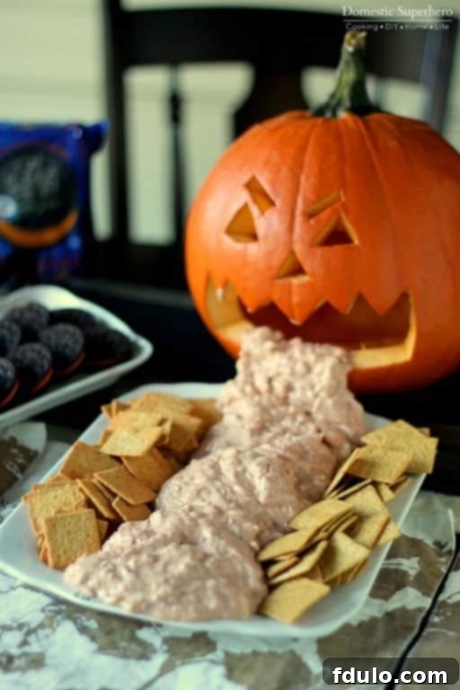 A festive Halloween Chunky Cheese Dip served in a carved pumpkin made to look like it's vomiting, surrounded by crackers.