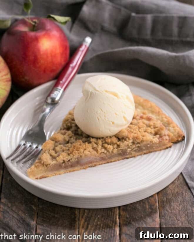 Apple Pie Pizza - A dessert pizza made with a pastry crust, spiced apples, and a crumb topping! An easy apple recipe with the taste of a Dutch Apple Pie! A delectable slice of Apple Pie Pizza, perfectly golden, with a scoop of vanilla ice cream melting on top, presented on a pristine white dessert plate. The blurred background shows the rest of the pizza, hinting at more deliciousness.