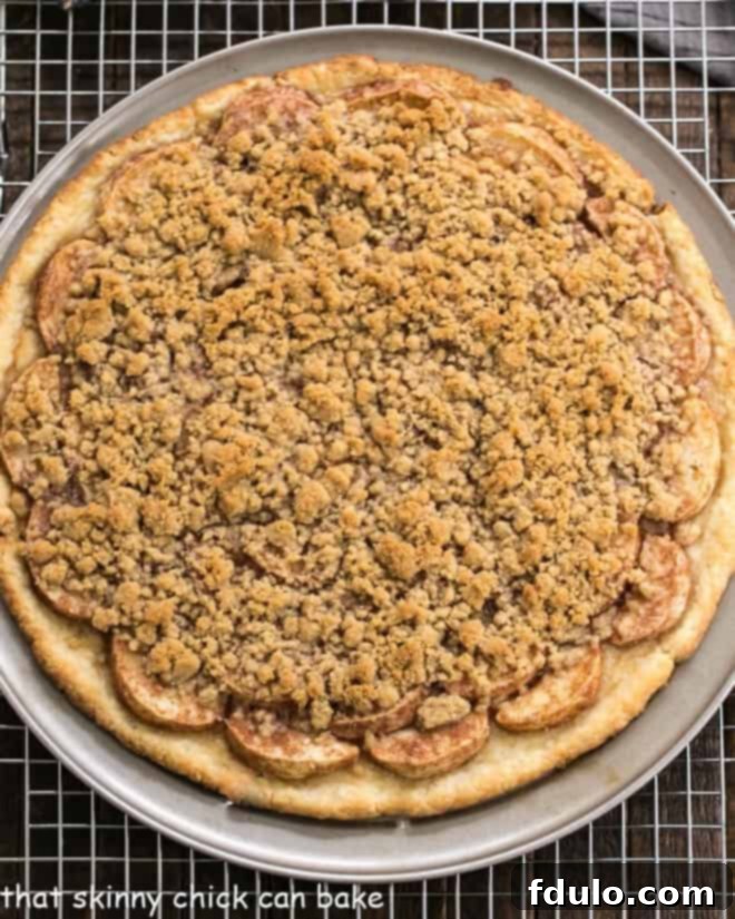 Apple Pie Pizza - A dessert pizza made with a pastry crust, spiced apples, and a crumb topping! An easy apple recipe with the taste of a Dutch Apple Pie! An inviting overhead shot of a freshly baked Apple Pie Pizza, golden brown and glistening with its spiced apple filling and crumb topping, resting on a rustic pizza pan, ready to be sliced and served.