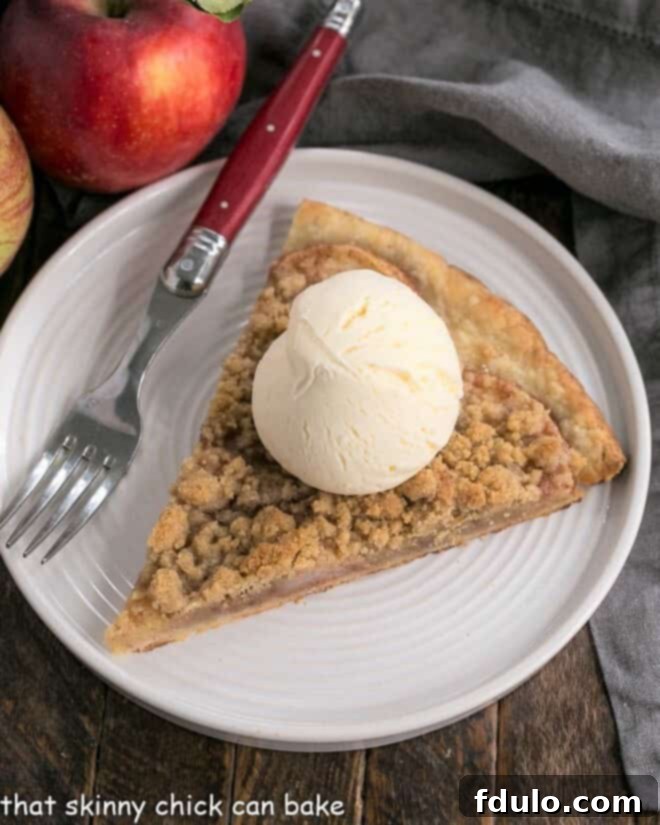 Apple Pie Pizza - A dessert pizza made with a pastry crust, spiced apples, and a crumb topping! An easy apple recipe with the taste of a Dutch Apple Pie! A close-up shot of a single slice of Apple Pie Pizza, richly topped with a melting scoop of vanilla ice cream, served on a elegant white plate. The golden crust and spiced apples are clearly visible, promising a delightful treat.