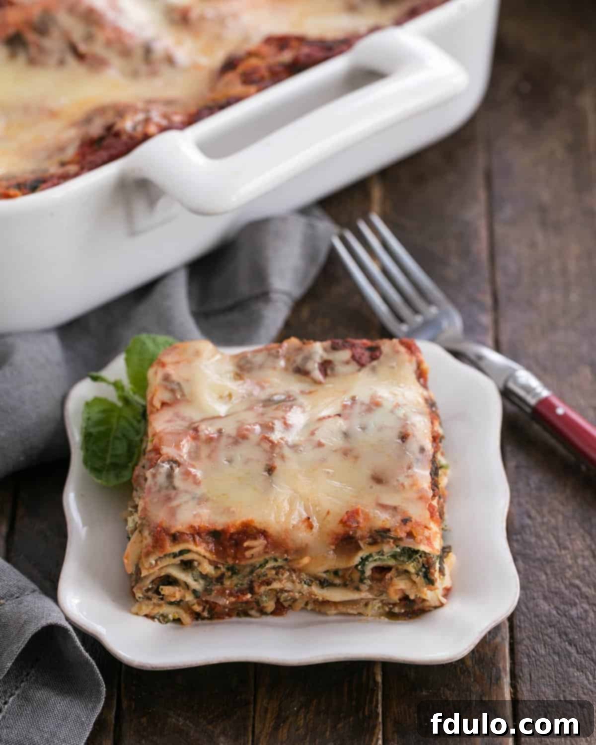 A delectable slice of Easy Spinach Lasagna, showing off its rich, layered interior, is presented on a pristine white plate, with the rest of the golden-brown casserole dish visible in the background, ready for more servings.