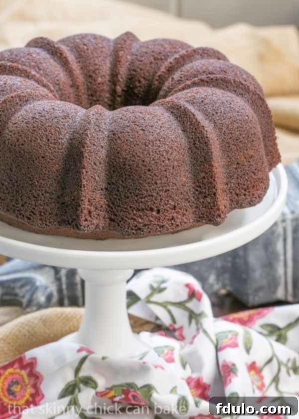 A beautifully baked Chocolate Buttermilk Bundt Cake sits on a white ceramic cake stand, accompanied by a charming floral napkin, ready to be served.