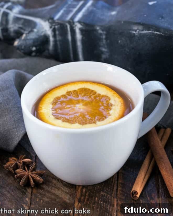 Hot Apple Cider Recipe flavored with vanilla, oranges and warm autumn spices. Perfect for holiday parties, apres ski parties or anytime you need to warm your soul A round white mug filled with hot Mulled Apple Cider, garnished with a vibrant orange slice and a cinnamon stick, inviting a moment of warmth.