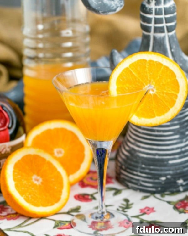 The Grand Mimosa 7 Grand Marnier Mimosas in a glass pitcher with a glass garnished with an orange slice and orange halves in the foreground