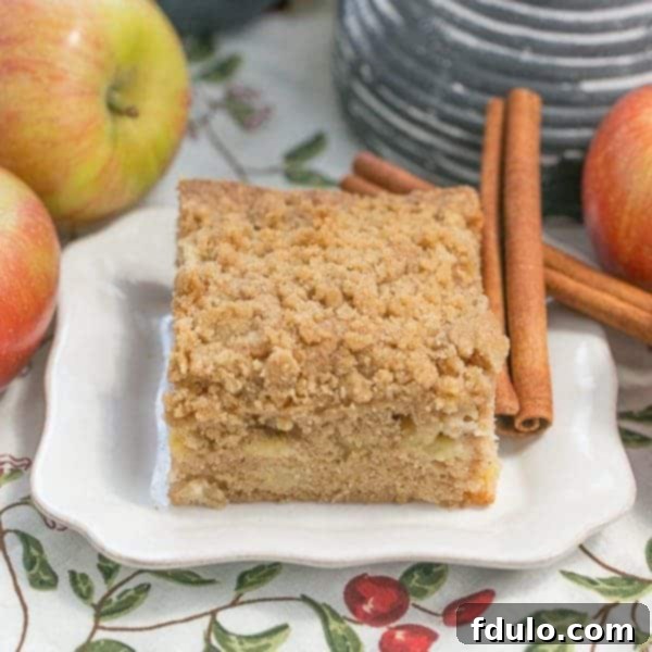Apple Coffee Cake slice on a white plate surrounded by fresh apples and cinnamon sticks