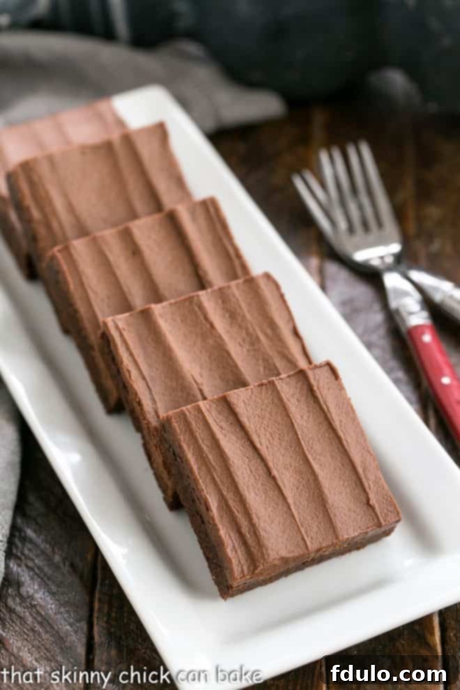 Irresistibly Chewy Cocoa Brownies with a decadent, extra-thick layer of chocolate buttercream frosting! Line of luscious Chewy Cocoa Brownies on a white ceramic tray, each perfectly frosted, with two red-handled forks poised for enjoyment.