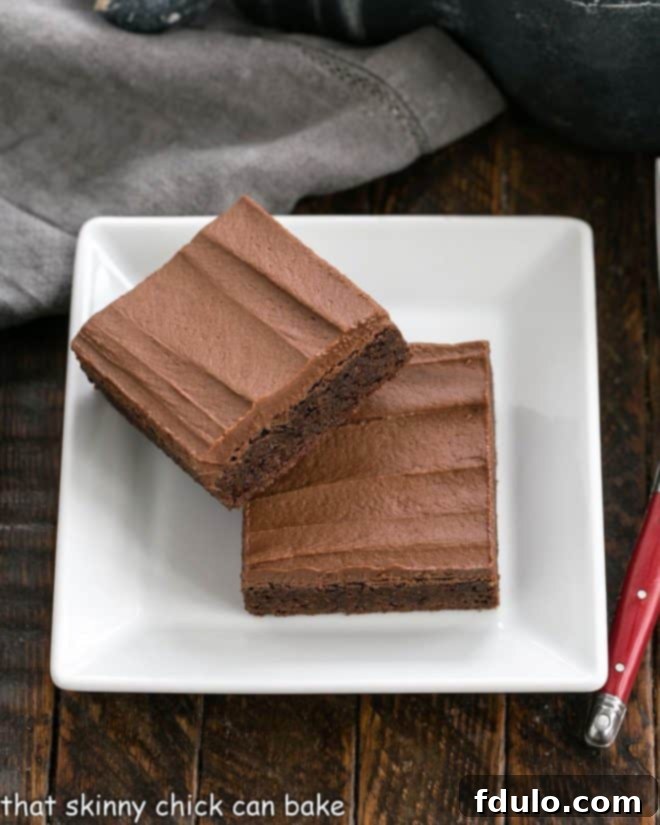 Decadent Chewy Cocoa Brownies crowned with an exquisitely thick layer of luscious chocolate buttercream! Two perfect squares of Chewy Cocoa Brownies, generously frosted, presented on a pristine white plate, ready to be enjoyed.