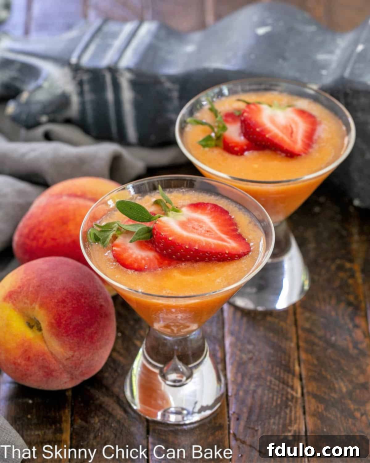 Peach Bellini Cocktail - A slushy version of the famous Italian champagne cocktail Two vibrant Peach Bellinis, garnished with fresh strawberry slices, sitting on a rustic wooden table.
