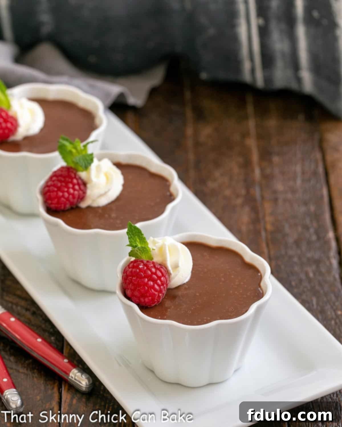 Three individual Irish cream pots de creme resting on a white ceramic tray, accompanied by delicate red-handled spoons. Each custard is topped with whipped cream and a fresh raspberry, highlighting its readiness for enjoyment.