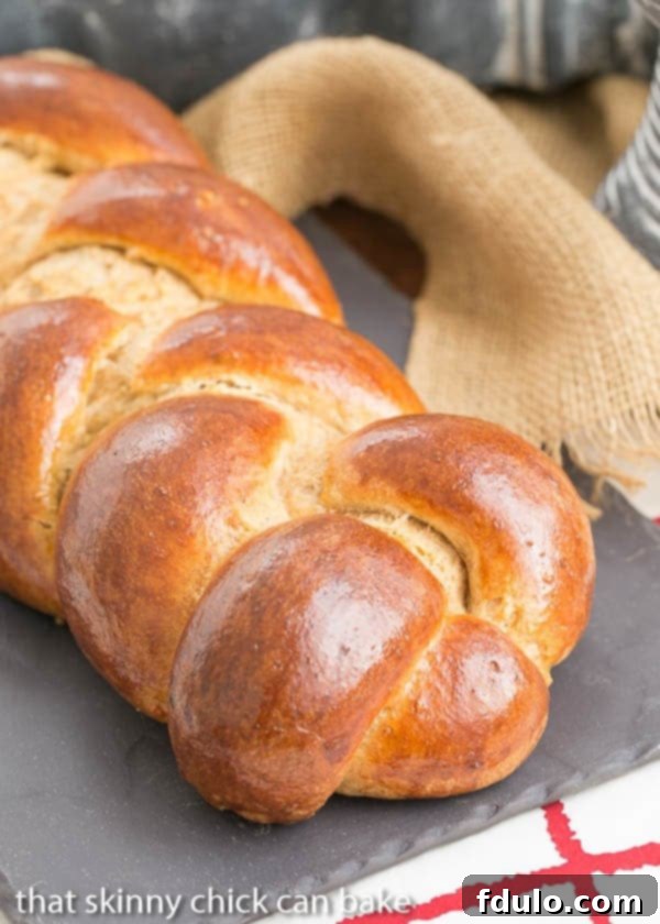 A perfectly braided loaf of Whole Wheat Challah, golden brown and glistening, resting on a slate tray.