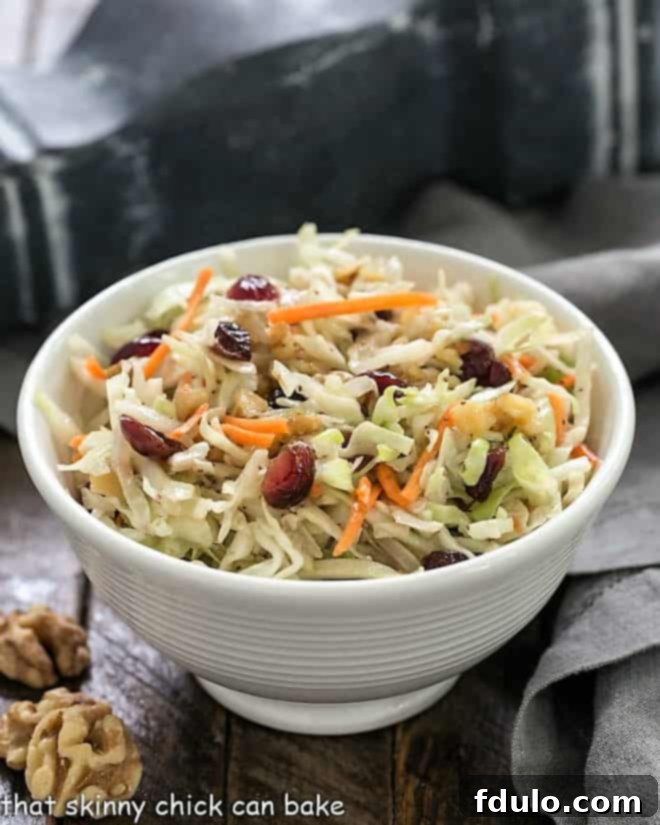 This Easy No-Mayo Coleslaw recipe can be made in minutes. Dotted with vibrant dried cranberries and crunchy toasted walnuts, it's a crowd-pleasing side dish perfect for cookouts, picnics, and potluck dinners without the worry of mayonnaise. Easy No-Mayo Coleslaw recipe presented in a small white ceramic bowl, accompanied by a soft gray napkin and whole walnut halves for garnish.