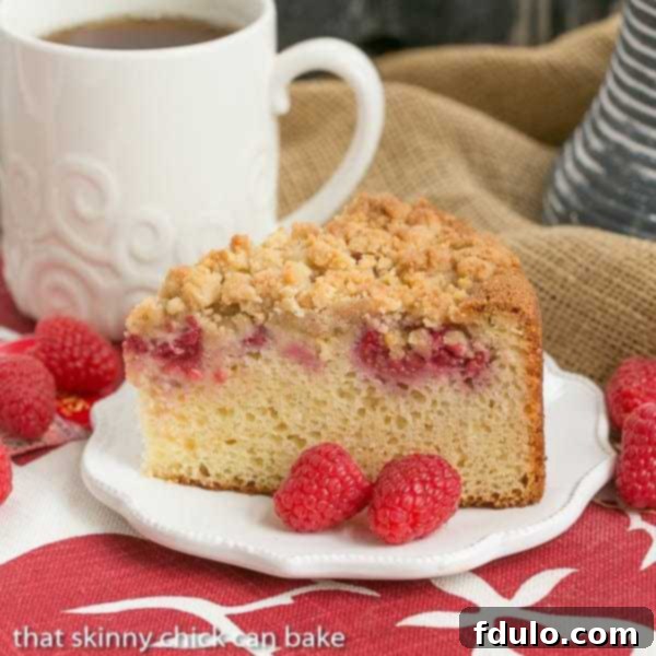 A slice of moist raspberry coffee cake on a white plate, garnished with fresh raspberries, showcasing its crumb topping and berry-filled interior.