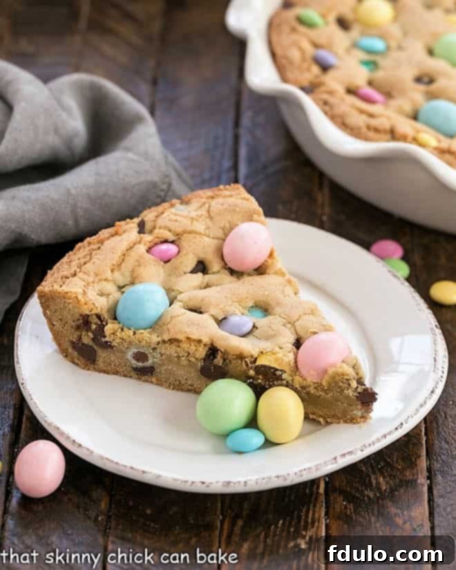 Easter Cookie Cake - A giant, chewy cookie packed full of chocolate chips and Easter candy A perfectly sliced piece of Easter cookie cake, rich with chocolate chips and colorful Easter candies, resting on a white plate. The rest of the large cookie cake, studded with more candy, sits invitingly in the background.
